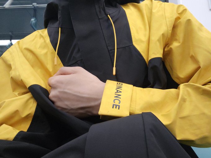 A person wearing a yellow and black "BINANCE" jacket stands with arms crossed, suggesting they were a crypto/finance industry professional attending an event at a fancy venue. The image captures the "standing for over an hour" experience mentioned in the post, showcasing the formal yet waiting aspect of trying to access an exclusive meeting.
