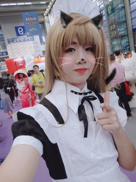 A cosplayer dressed in a black and white French maid outfit with cat ears, whiskers, and a painted cat nose, taking a selfie while making a peace sign at a convention.
