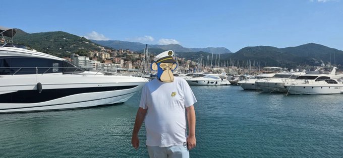 A person wearing a Bored Ape Yacht Club mask stands in front of a harbor filled with yachts, embodying the luxury associated with the NFT collection. The image features a prominent white yacht in the foreground and a coastal town in the background.
