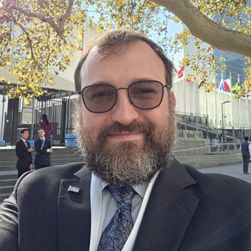 A bearded man wearing glasses, a dark suit, and patterned tie smiles for a selfie outside a building displaying the iconic UN logo and international flags. The image relates to the post by featuring Charles Hoskinson, the founder of Cardano ($ADA), at the United Nations headquarters.