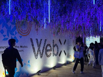 An illuminated 'WebX' event entrance decorated with hanging purple wisteria-like flowers and neon vertical lights at night.