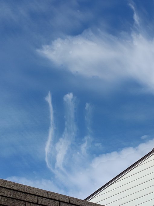 A photograph of a blue sky with wispy clouds that appear to form the shape of a hand with fingers pointing upward, seen above a roofline.