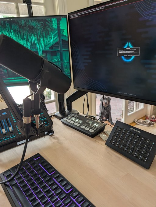 A desktop streaming setup featuring dual monitors, a professional microphone, Stream Deck, Elgato Stream Deck, and an analog keyboard with a dog visible in the background. The setup supports the post's reference to a "hacked" desktop and includes a Stream Deck ("clicky thangies") for stream control.