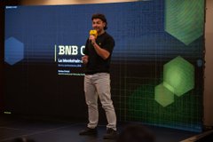 A man in a dark t-shirt and light gray pants stands on a stage speaking into a microphone against a digital backdrop displaying the logo 'BNB' and a green honeycomb pattern.