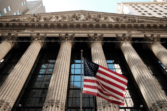 A low-angle photograph of the New York Stock Exchange building, featuring its grand Corinthian columns, classical pediment sculptures, and a large American flag hanging prominently in the center.