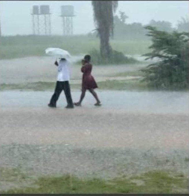 Two people wade through a flooded road in heavy rain, one holding an umbrella, as they brave the storm together.