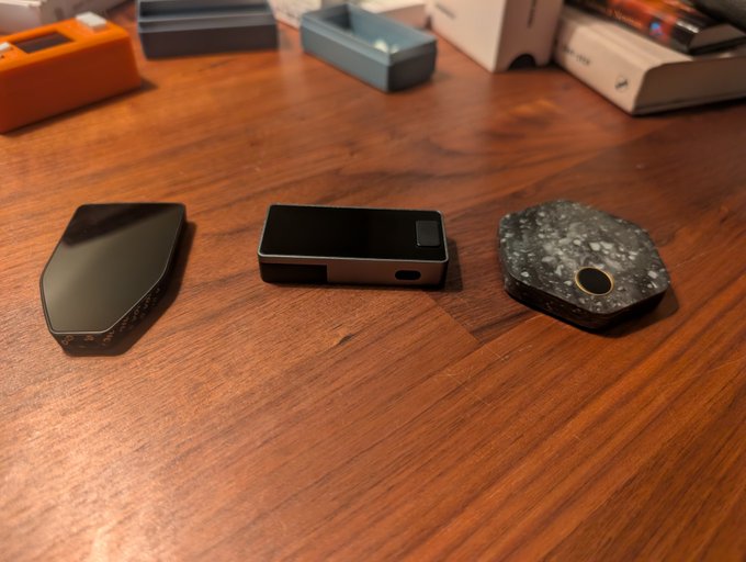 Three electronic vaping devices arranged on a wooden table, with various device boxes and accessories visible in the background.