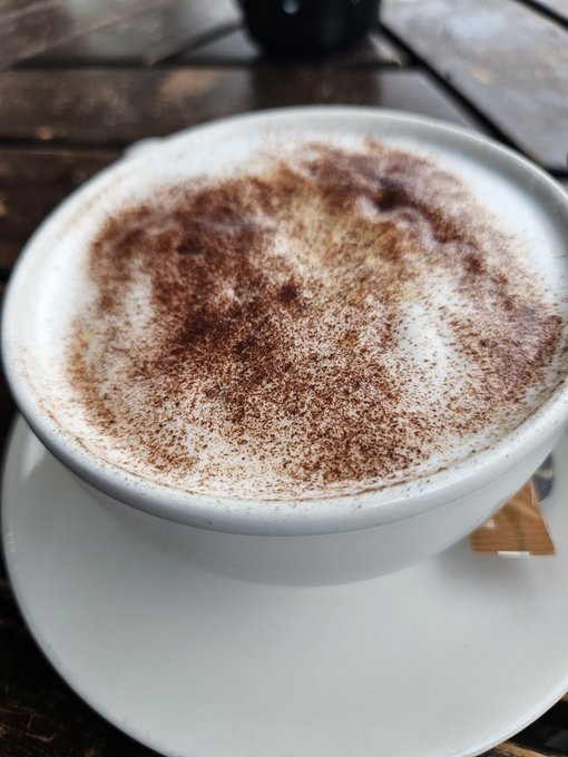 A close-up shot shows a white ceramic cup filled with a frothy beverage, possibly coffee or hot chocolate, generously dusted with cocoa powder, sitting on a saucer on a dark wooden table.