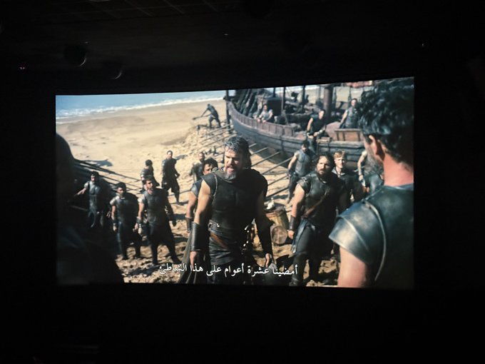 A photo of a movie theater screen showing a scene from *Troy* (2004), featuring Gerard Butler as King Leonidas surrounded by warriors on a beach, with Arabic subtitles at the bottom. The "odyssey" reference ties in with the epic, journey-like setting of the film being viewed in theaters.