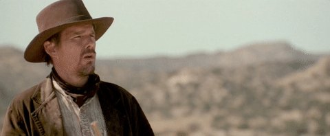 The image features a man wearing a cowboy hat, standing in an open area with mountains in the background. The man appears to be dressed as a cowboy or a rancher, possibly from the Wild West era. The scene suggests that he might be taking a break or waiting for someone while being surrounded by nature and the picturesque landscape of the mountains.