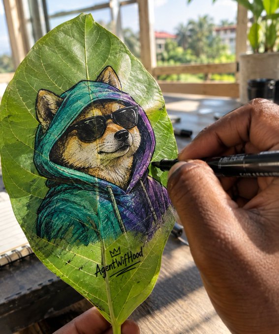An artist's hand uses a black marker to draw a detailed portrait of a Shiba Inu dog wearing sunglasses and a colorful teal and purple hoodie onto a large green leaf, signed 'AgentWiiHood' with a small crown.