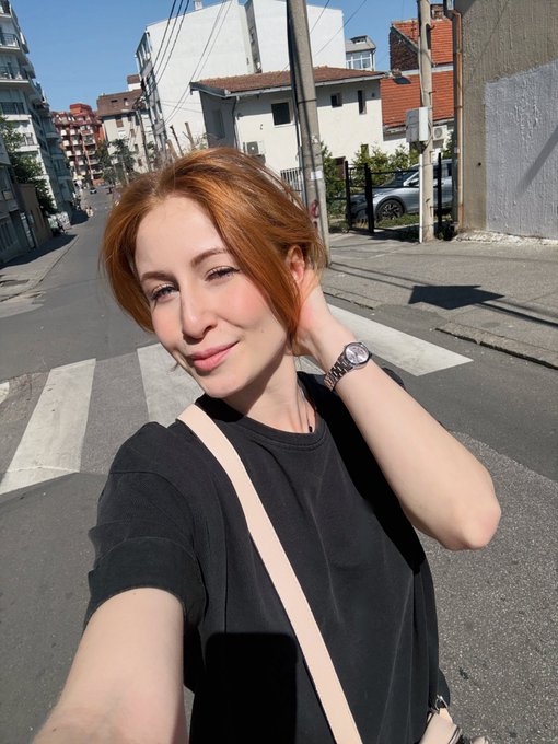 A red-haired woman wearing a black t-shirt takes a selfie on a sunny street corner with a crosswalk, with houses and a parked car visible in the background.