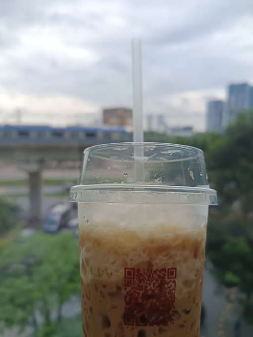 A close-up of a plastic cup containing an iced coffee/bubble tea drink with a QR code on it, with an elevated train and city skyline blurred in the background.