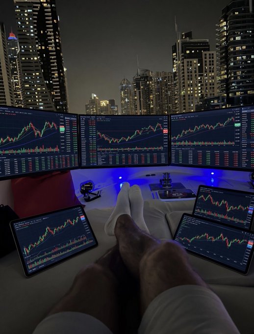 A first-person view of a person relaxing in socks with their feet up, watching financial candlestick charts across multiple monitors and tablets displaying green and red price movements, set against a dramatic nighttime city skyline—all humorously underscoring the contrast between the small $130 trading portfolio and the high-tech, professional-looking setup.