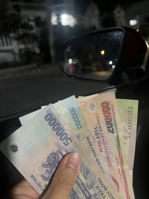 A hand holds several Vietnamese Dong banknotes (50,000 and 200,000 denominations) inside a car at night, with the side mirror and street lights visible through the window.
