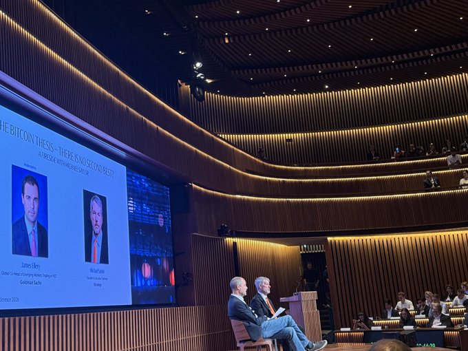 Two men sit on a stage in a modern, tiered auditorium at the Goldman Sachs Digital Assets Conference 2026, with a large screen behind them displaying "THE BITCOIN THESIS - THERE IS NO SECOND BEST: A DEBATE WITH MICHAEL SAYLOR" featuring headshots of James Luby and Michael Saylor. The image supports the post's message about crypto industry momentum, visually capturing the well-attended Goldman Sachs event where major trading desks are reportedly seeing furious onboarding of new digital asset clients.