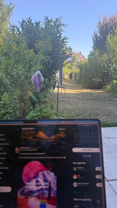 An outdoor scene with a laptop screen displaying social media feeds in the foreground, with a garden featuring a purple decorative stake and a path in the background.