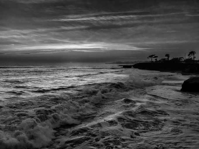 A black and white photograph of ocean waves crashing along a coastline under a cloudy, overcast sky.