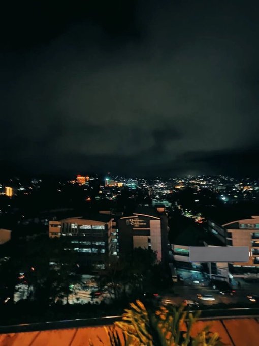 The image shows a city skyline at night with buildings lit up against the dark sky. The scene captures the beauty of the city lights as they illuminate the surrounding area. The photo appears to have been taken from an elevated position, possibly from a rooftop or balcony, providing a unique perspective on the cityscape below.