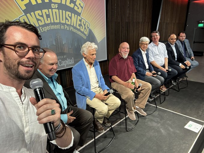 A group selfie of six men seated on stools on a stage at what appears to be a 'Physics of Consciousness' event, with a banner in the background partially reading 'An Experiment in Psi Phenomenon' and 'HOSTED BY Capital'.
