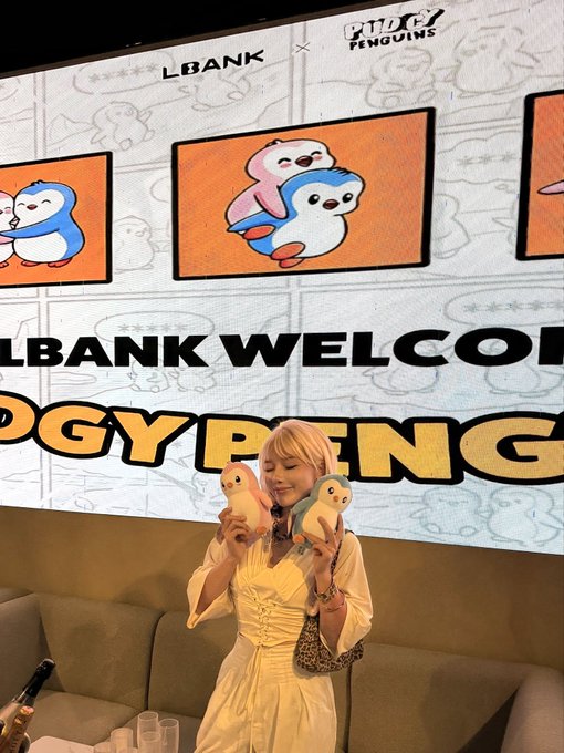 A blonde woman in a cream-colored dress holding two Pudgy Penguins plush toys at an event with a backdrop reading 'LBANK WELCOMES PUDGY PENGUINS' featuring cartoon penguin characters.