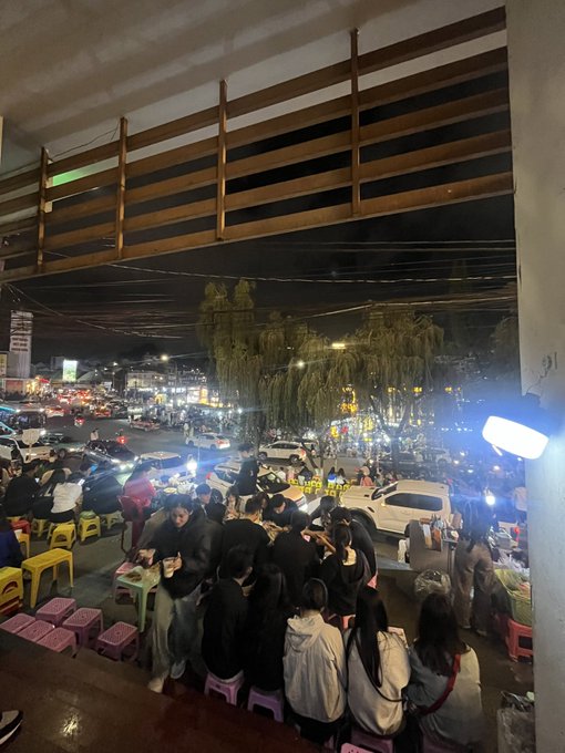 Nighttime photo taken from an elevated vantage point showing a crowd of people gathered on small colorful plastic stools around low tables, possibly at a street food or dining area.