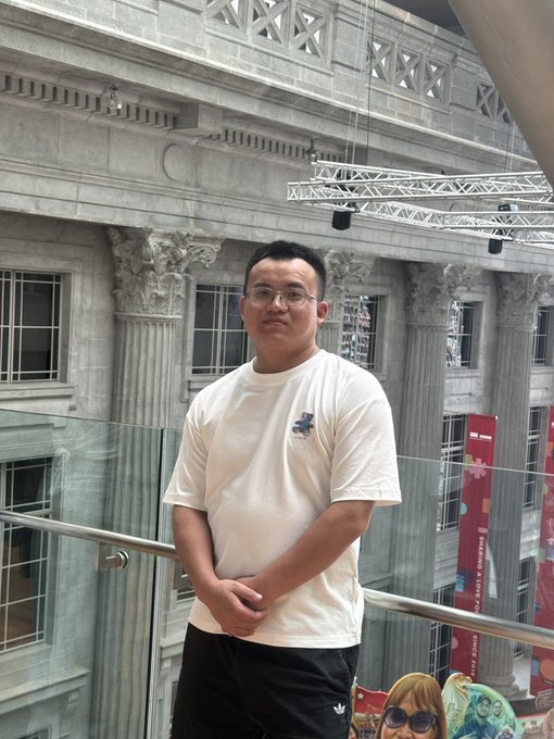 A man wearing glasses and a white t-shirt posing in front of an ornate building with classical columns.