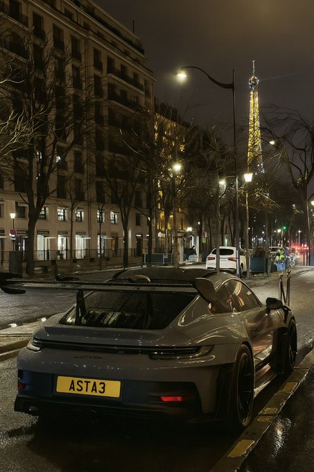 A gray Porsche sports car with license plate 'ASTA3' parked on a Parisian street at night, with the illuminated Eiffel Tower visible in the distance.