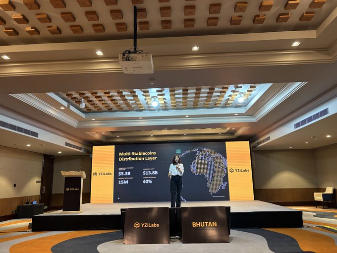 A conference stage with a presenter standing before a large screen displaying "Multi-Stablecoin Distribution Layer" stats ($5.3B abs, $13.8B, 15M, 40%) for YZiLabs, with "BHUTAN" signs in front.