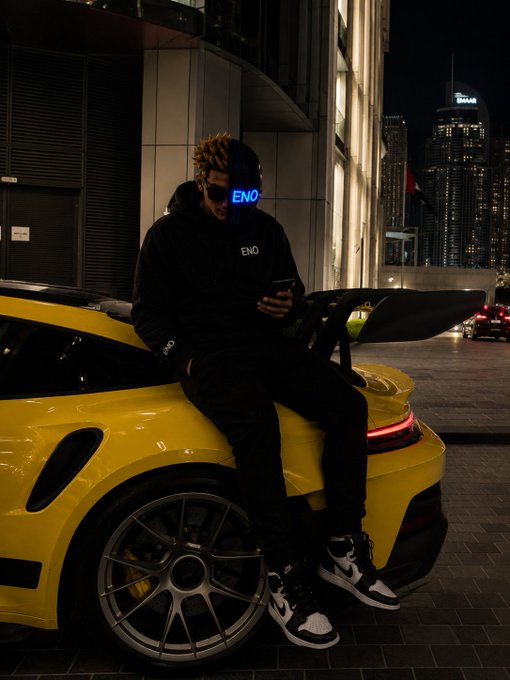 A person sits on the back of a yellow sports car at night in the city, wearing a black hoodie with "ENO" branding, looking at their phone. The image complements the post's "Good night" crypto message by capturing a moment of rest amid the urban hustle, symbolizing taking a break before tomorrow's market activity.