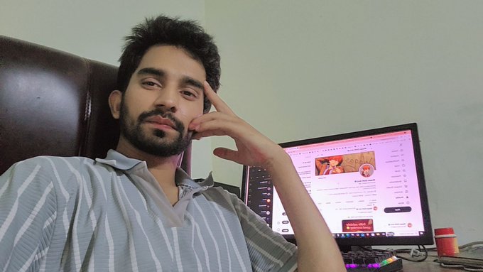 A young bearded man sits at a desk with a computer monitor, resting his hand near his face.