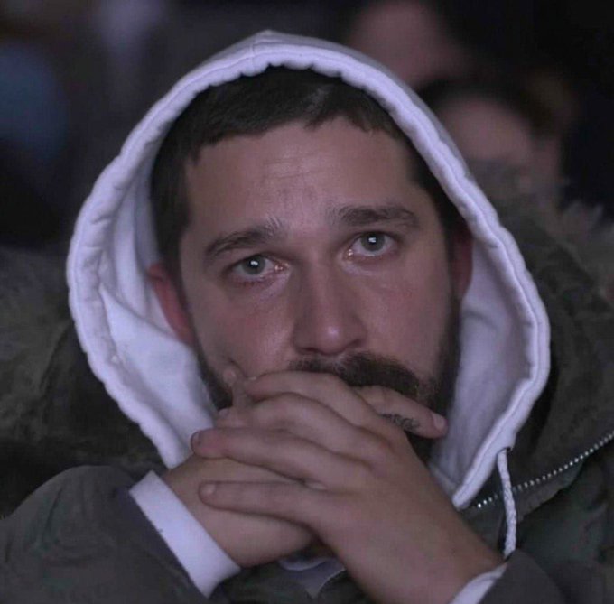 A close-up portrait of a bearded man wearing a white hoodie, looking emotional with his hands clasped over his mouth.