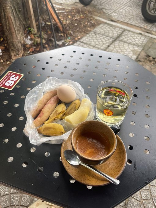 A black perforated café table set with a Vietnamese coffee in a brown ceramic cup, a glass of water/tea, and a plate of sweet potatoes and banana, with a 'Cộng' sticker visible.