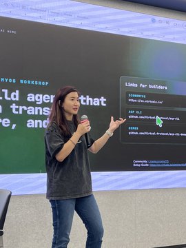 A woman in a grey t-shirt and jeans holds a microphone while presenting in front of a large screen displaying code and the text 'build agents that think, transfer, and learn'.