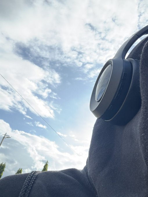 A first-person view looking up at a pair of black over-ear headphones resting on a person's lap against a partly cloudy blue sky.
