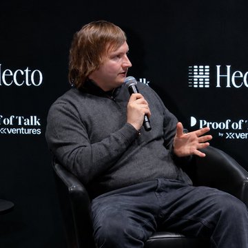 In the image, a man with long hair is sitting in a chair while holding a microphone. He appears to be speaking into it or possibly conducting an interview. The background features a black wall with white text that reads "INSIGHT: @aave founder @StaniKulechov says, 'The bigger dream is that Aave becomes embedded infrastructure for finance.'" This suggests that the man might be discussing the founder of a company called Insight, which could potentially be related to the financial industry.