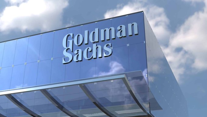 The top corner of a modern glass building displays the 'Goldman Sachs' logo in white lettering against a reflective blue facade, set against a partly cloudy sky.