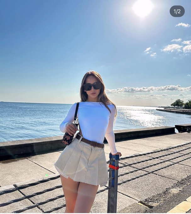 The image features a woman standing next to a body of water, wearing a white top and skirt. She has her handbag over her shoulder and sunglasses on her head. The background includes a beach setting with an ocean view. The caption for the image reads "happy sunday CT a little family time today still making content about @quipnetwork and @RiverdotInc what'S everyone up to today?".