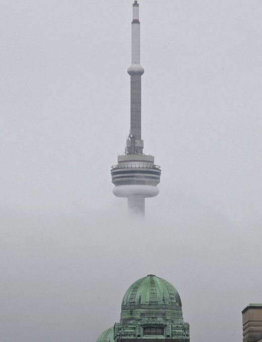 A photograph of the upper section of the CN Tower in Toronto emerging from heavy fog, with the green domed roof of a nearby building visible at the bottom.