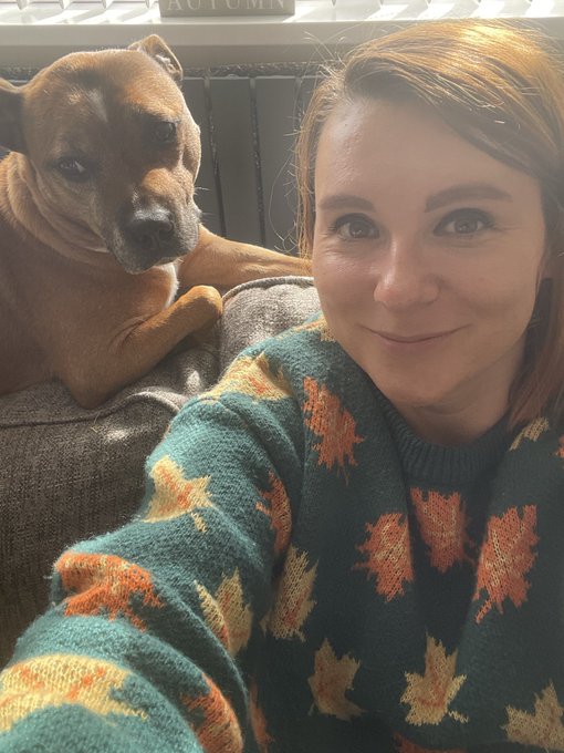 A woman wearing a cozy green sweater with orange and yellow maple leaf patterns poses for a selfie alongside a brown dog resting on a couch, with the partial word "AUTUMN" visible at the top of the frame. The fall-themed sweater and "Gm" post combine for a warm, seasonal morning greeting.