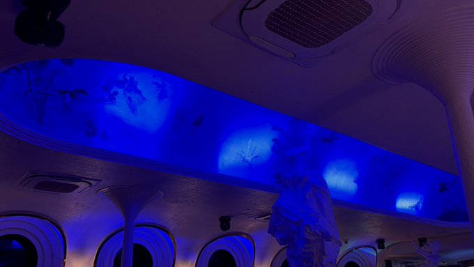 A dimly lit cabin interior bathed in deep blue lighting features an elongated ceiling panel displaying a world map projection, with a costumed figure standing beneath it near what appears to be a futuristic or themed vessel's porthole-style windows. The atmospheric, dreamlike setting with its globe motif visually echoes the post's message about long-term commitment and perseverance in cryptocurrency, suggesting a journey or voyage spanning the world that requires dedication before yielding results.