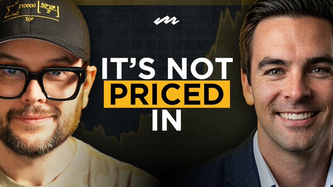 A thumbnail image for a video titled 'IT'S NOT PRICED IN' featuring two men, one wearing a cap with '210000' text and glasses, and another man in a suit smiling, against a dark stock chart background.