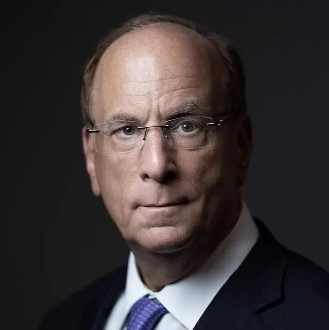 A professional headshot of an older, balding man wearing glasses, a dark suit, and a purple tie against a dark background.