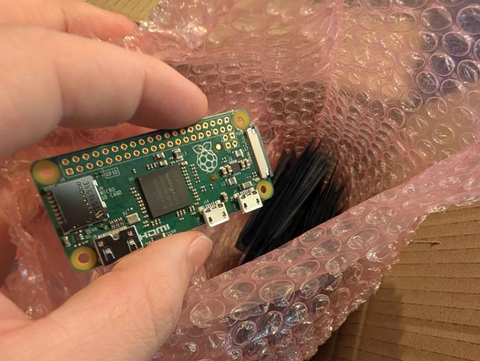 A hand holds a green Raspberry Pi Zero circuit board over a cardboard box containing pink bubble wrap and a bundle of black components.