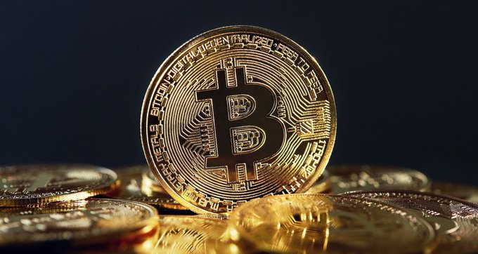 A close-up photograph of a physical Bitcoin token standing upright on a pile of other gold-colored coins against a dark background.