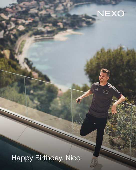 Nico Hulkenberg laughs joyfully on a terrace overlooking a stunning Mediterranean coastline, with "NEXO" and "Happy Birthday, Nico" text framing the celebratory post wishing the F1 driver a long, leisurely birthday.