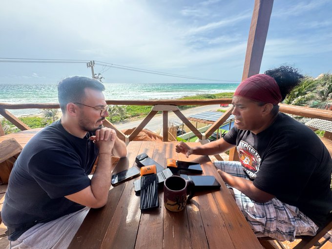The image shows two men sitting at a wooden table, engaged in a conversation while enjoying their drinks. One of them has a cup in front of him, possibly containing coffee or another beverage. The scene takes place outdoors, with a view of the ocean visible through the window behind the men. The post seems to be promoting Bitcoin and cryptocurrency, as indicated by the use of the term "bitcoin cafe" and the reference to the bear market.