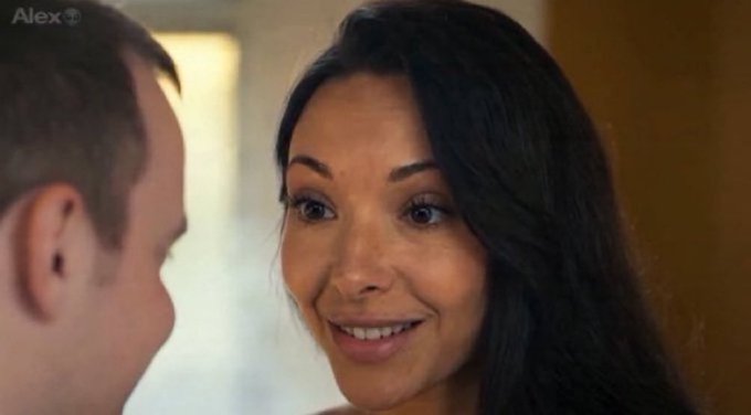A screenshot from a video showing a close-up of a smiling woman with long dark hair facing a man, with the watermark 'Alex' visible in the top left corner.