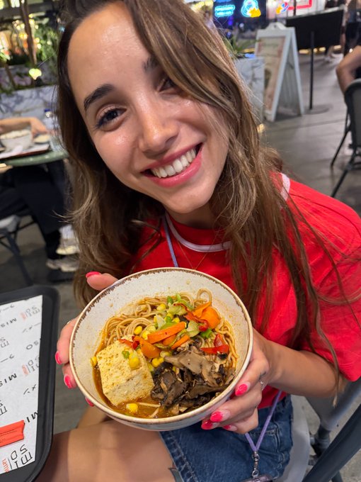 A smiling woman in a red shirt holds a bowl of noodles with vegetables, mushrooms, and tofu at what appears to be a food market or restaurant.