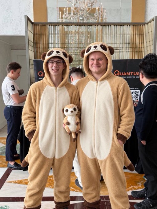 Two men wearing matching tan meerkat/kinkajou onesie costumes standing together indoors at what appears to be an event with a 'QUANTUS' banner in the background.